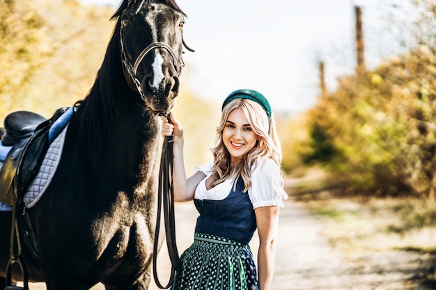 Retty blonde en robe traditionnelle marche avec grand cheval noir à l'extérieur