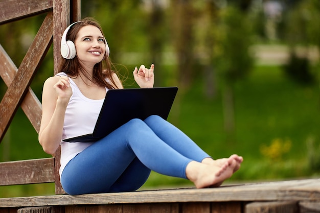 Réunion en ligne dans la nature par une femme joyeuse dans un casque sans fil avec un ordinateur portable à l'extérieur