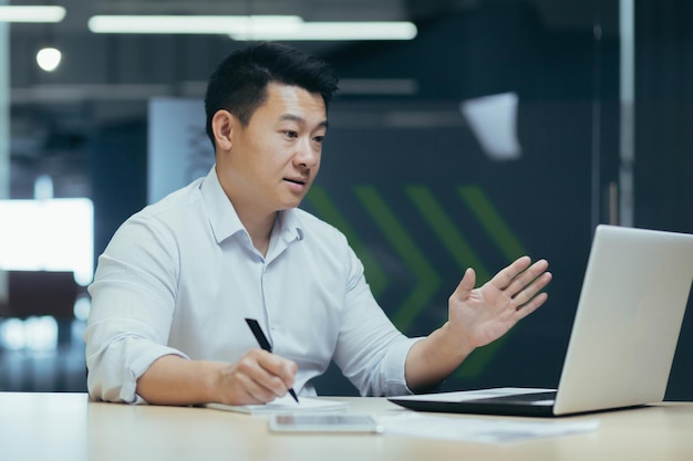 Réunion en ligne d'un homme d'affaires asiatique regardant l'écran d'un ordinateur portable et parlant avec des collègues enregistrant des données