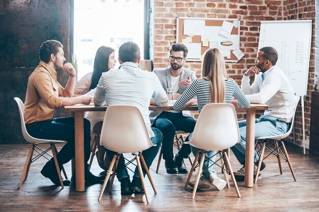 Réunion en matinée. Groupe de six jeunes discutant de quelque chose assis à la table au bureau ensemble