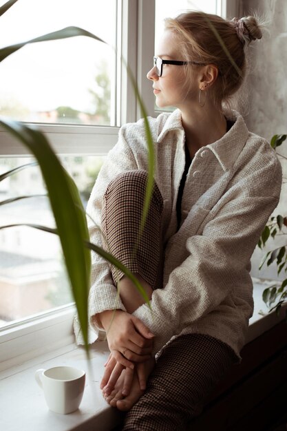 Rêve vertical femme blonde réfléchie tranquille dans des verres et un pull chaud assis sur le rebord de la fenêtre avec une tasse de café