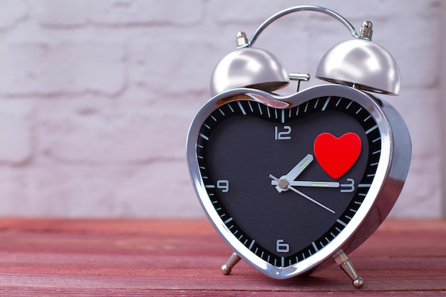 Réveil en acier sous forme de coeur avec un coeur rouge sur une table en bois rose et avec un mur de briques blanches Saint Valentin