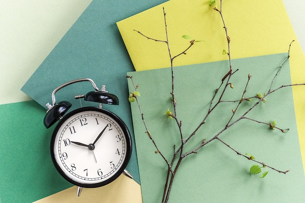 Réveil noir et branches d'arbre avec de jeunes feuilles en fleurs sur fond de nuances vertes géométriques.