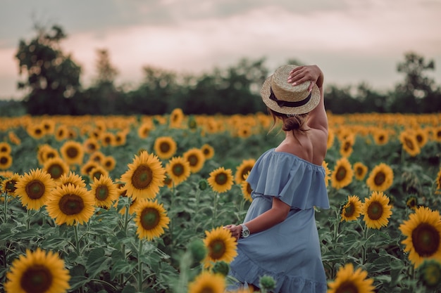 Rêver de jeune femme en robe bleue tenant un chapeau avec une main dans un champ de tournesols à l'été, vue de son dos. Avoir hâte de. copie espace