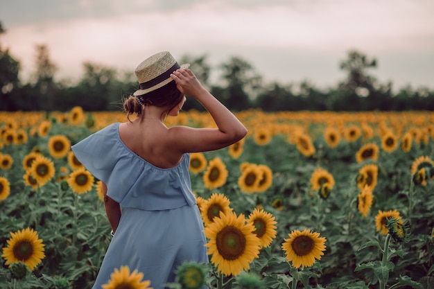 Rêver de jeune femme en robe bleue tenant un chapeau avec une main et s'éloignant dans un champ de tournesols en été, vue de son dos. Regardant sur le côté. copie espace