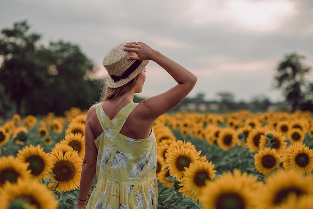Rêver de jeune femme en robe jaune tenant un chapeau avec une main et s'éloignant dans un champ de tournesols en été, vue de son dos. Regardant sur le côté. copie espace