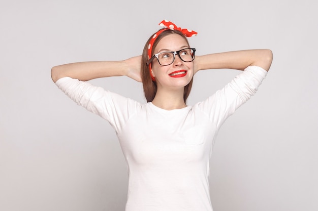 Rêver d'un portrait plein d'espoir d'une belle jeune femme émotive en t-shirt blanc avec des taches de rousseur, des lunettes noires, des lèvres rouges et un bandeau. tourné en studio intérieur, isolé sur fond gris clair.