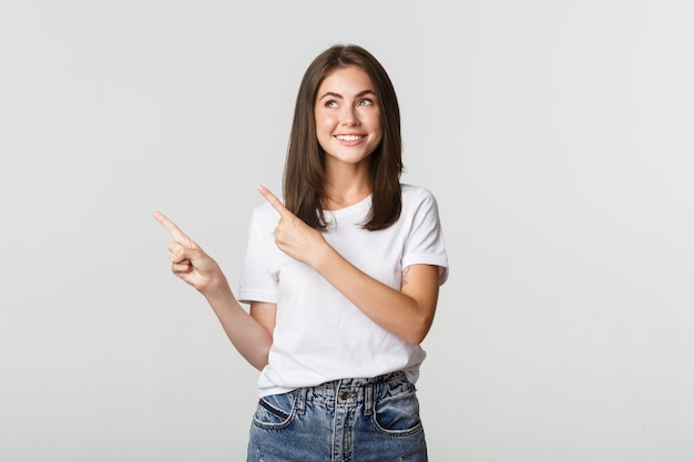 Rêveuse Belle Fille Souriante, Pointant Du Doigt Le Coin Supérieur Gauche Et Regardant Heureux Au Logo.