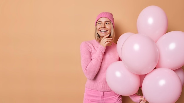 Rêveuse jeune femme charmante organise une fête d'adieu touche la mâchoire regarde doucement ailleurs a la bonne humeur tient un tas de ballons à l'hélium pose sur fond marron espace de copie vierge pour la promotion