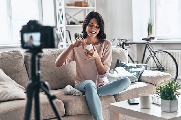 Revue de la crème lissante. Belle jeune femme testant un produit de beauté et souriant tout en faisant une vidéo sur les réseaux sociaux