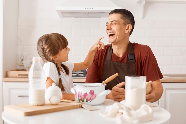 Riant jeune adulte aux cheveux noirs portant un t-shirt marron et un tablier cuisinant dans la cuisine avec sa petite fille, un enfant a enduit le nez du père de farine, exprimant des émotions heureuses.