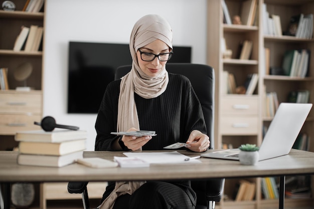 Riche femme d'affaires musulmane travaillant sur un ordinateur portable comptant de l'argent en espèces calculer le bénéfice du revenu