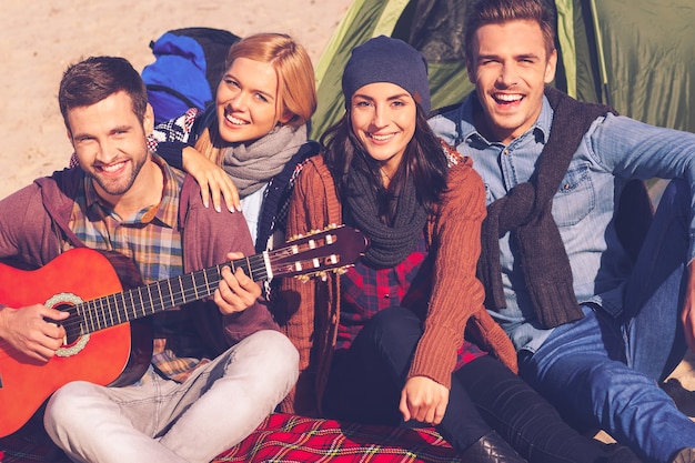 Rien que les meilleurs amis. Vue de dessus de quatre jeunes gens heureux assis près de la tente ensemble tandis que jeune bel homme jouant de la guitare et souriant