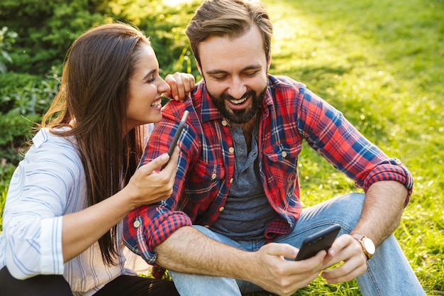 rire couple homme et femme vêtus de vêtements décontractés utilisant un téléphone portable tout en se reposant dans un parc verdoyant