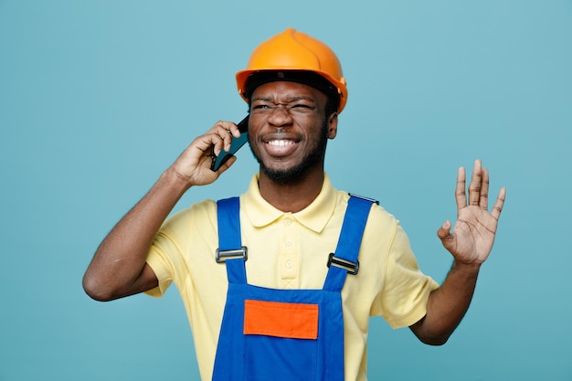 Rire jeune constructeur afro-américain en uniforme parle au téléphone isolé sur fond bleu