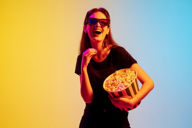 Rire. Jeune femme caucasienne avec bouquet de pop-corn sur fond de studio dégradé bleu-jaune en néon. Concept de film, émotions humaines, expression faciale, ventes, publicité. Regarder du cinéma avec des lunettes.