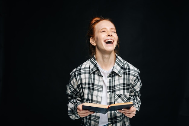 Rire jeune femme étudiante avec les yeux fermés tenant un livre ouvert sur fond noir isolé Joli modèle de dame rousse montrant émotionnellement les expressions faciales dans la surface de studio