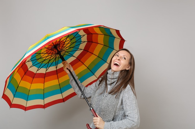 Rire de jeune femme en pull gris, écharpe tenant un parapluie coloré isolé sur fond gris en studio. Gens de mode de vie sain émotions sincères, concept de saison froide. Maquette de l'espace de copie.