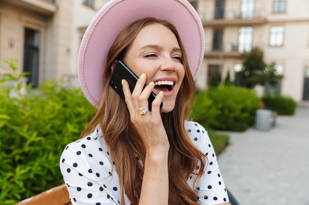 Rire jeune femme rousse s'asseoir sur un banc à l'extérieur dans la rue en parlant par téléphone mobile.