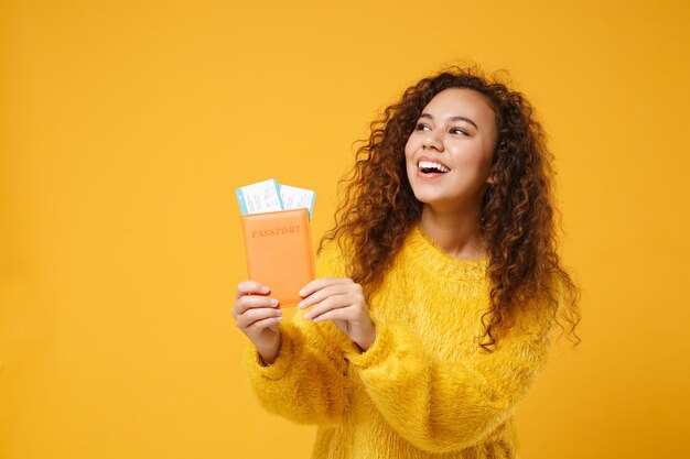 Rire jeune fille afro-américaine en pull de fourrure posant isolé sur fond de mur orange jaune portrait en studio. Concept de style de vie des gens. Maquette de l'espace de copie. Détenir des billets de carte d'embarquement de passeport.