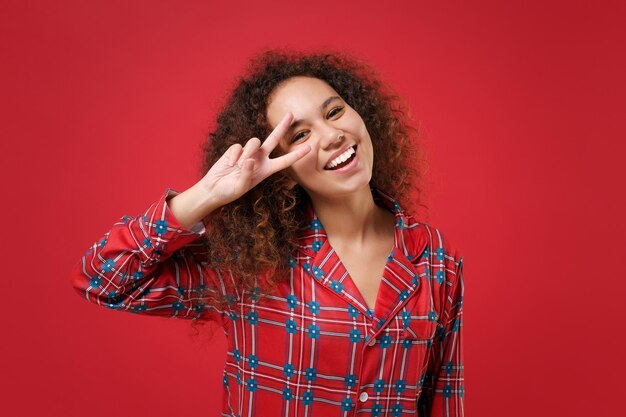 Rire jeune fille afro-américaine en pyjama homewear posant au repos à la maison isolée sur fond rouge portrait en studio. Détendez-vous le concept de mode de vie de bonne humeur. Maquette de l'espace de copie. Montrant le signe de la victoire.