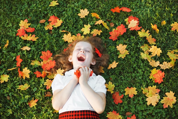 Rire petite fille couchée dans l&#39;herbe jette les feuilles d&#39;automne dans l&#39;air.