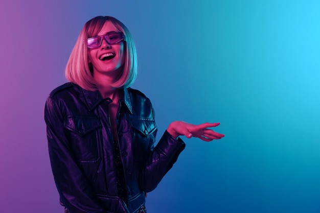 Rire sexy belle femme blonde dans une veste en cuir robe étincelante lunettes de soleil à la mode regardez de côté posant isolée dans le fond de la lumière de studio de couleur bleu rose fête au néon concept Cyberpunk copier l'espace