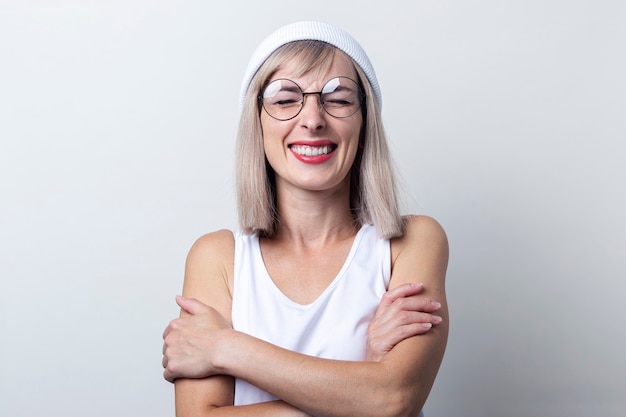 Rire les yeux fermés, la jeune femme tient les bras croisés dans un T-shirt blanc sur fond clair.