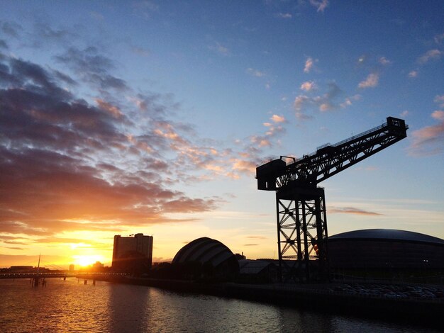 Photo rivière par des grues et des bâtiments contre un ciel nuageux vu à travers l'arc de clyde au coucher du soleil