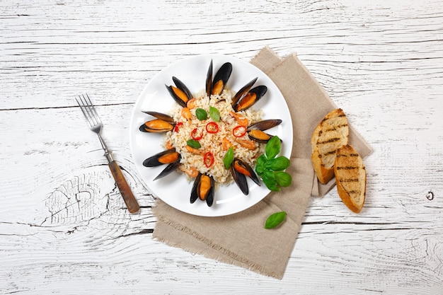 Riz frit aux moules de fruits de mer, crevettes et basilic dans une assiette avec toile de jute, baguette grillée et fourchette sur une table en bois craquelée blanche. Vue de dessus.