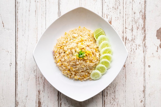 Photo riz frit savoureux avec des tranches d'œufs d'oignon de printemps oignon vert sur le dessus servir avec des tranches de concombre et de citron vert dans une assiette blanche sur fond de texture de bois blanc vue de dessus de table à plat