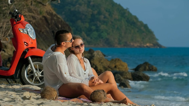 Road trip en scooter. Beau couple sur moto rouge en vêtements blancs sur la plage de sable. Les personnes juste mariées s'embrassent en marchant près des palmiers tropicaux, de la mer. Lune de miel de mariage au bord de l'océan. Location de moto.