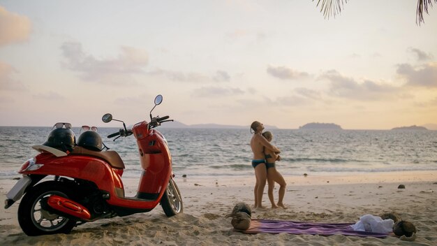 Road trip en scooter. Beau couple sur moto rouge en vêtements blancs sur la plage de sable. Les personnes juste mariées s'embrassent en marchant près des palmiers tropicaux, de la mer. Lune de miel de mariage au bord de l'océan. Location de moto.