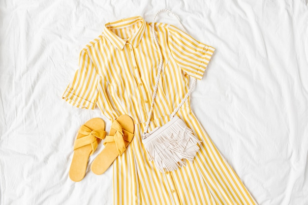 Robe jaune à rayures avec sac et chaussons sur lit blanc. Tenue d'été élégante pour femmes. Vêtements à la mode. Mise à plat, vue de dessus.