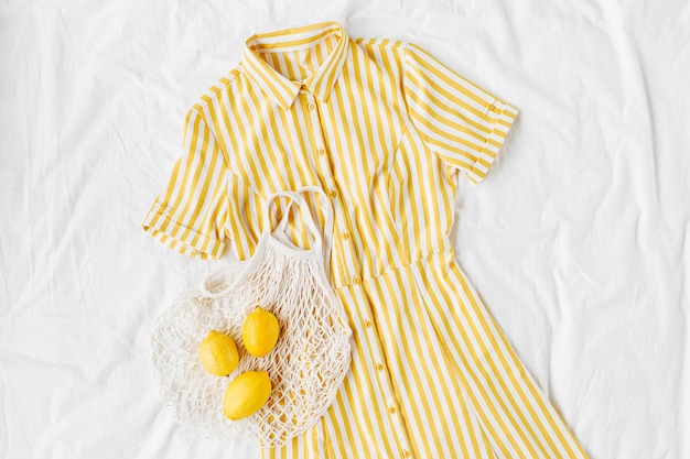 Robe jaune à rayures avec sac écologique et citrons sur lit blanc. Tenue d'été élégante pour femmes. Vêtements à la mode. Mise à plat, vue de dessus.