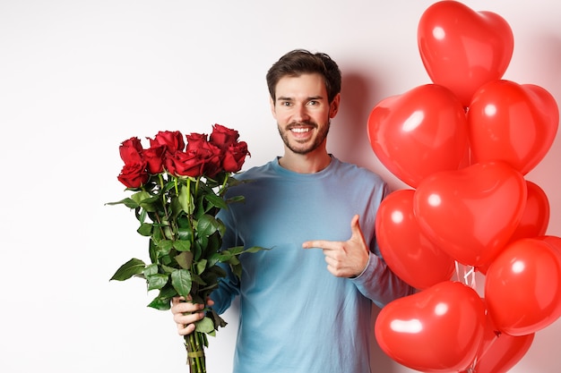 Romance de la Saint-Valentin. Heureux jeune homme avec des fleurs et des ballons coeur en rendez-vous avec son amant. Mec souriant pointant sur bouquet de roses rouges, debout sur fond blanc