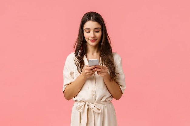 Romantique mignon, belle jeune femme en robe, discutant avec son petit ami, souriant, tenant le téléphone et tapotant l'écran pour commander en ligne, faire du shopping en utilisant l'application, debout mur rose