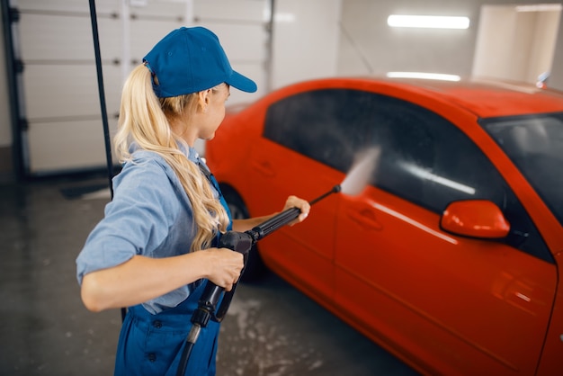 La rondelle femelle en uniforme nettoie l'automobile avec un pistolet haute pression dans les mains