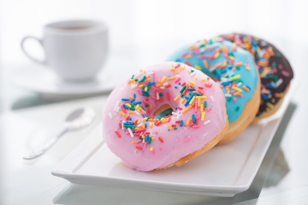 Rose, bleu et chocolat arrose des beignets sur une plaque blanche et une tasse de café