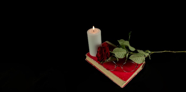 Une rose rouge avec un livre et une bougie allumée sur fond noir Du dessus d'une bougie allumée avec du rouge frais avec une rose sur un livre sur fond noir