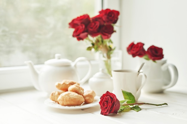 Roses rouges, thé et croissants sur une table près de la fenêtre, petit-déjeuner romantique pour la Saint-Valentin