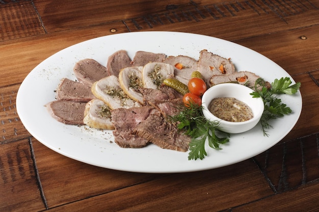 Rôti de boeuf cuit au four, rouleaux de poulet, langue et viande de porc avec légumes dans une assiette ovale blanche. Fond en bois.