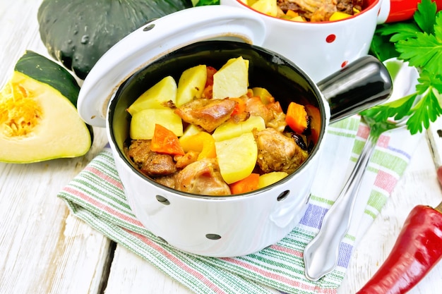 Rôti avec du poulet, des pommes de terre, de la courge et des poivrons dans un pot blanc à deux portions sur une cuillère à serviette persil sur le fond des planches en bois clair