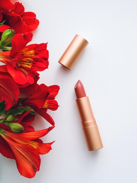 Rouge à lèvres nude dans un boîtier en métal doré et un bouquet d'alstroemeria rouge sur fond blanc