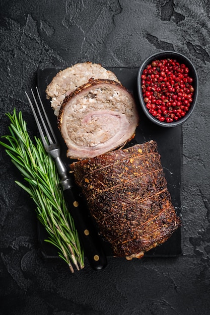 Roulade de porc au four Viande de porc roulée Porchetta sur une planche de marbre aux herbes Fond noir Vue de dessus