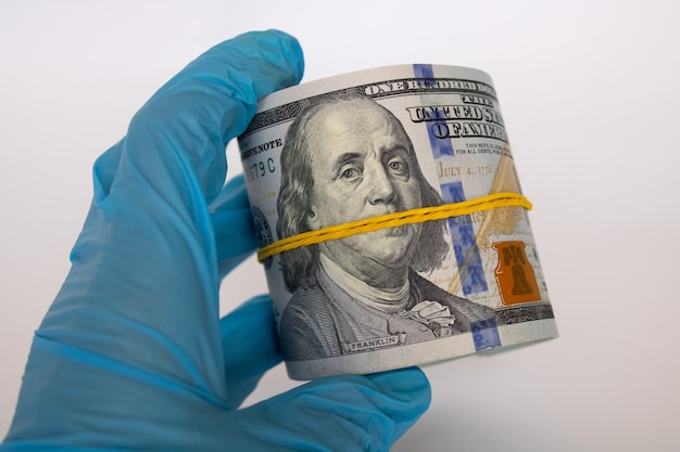 Rouleau de billets de cent dollars en main dans un gant médical sur fond blanc