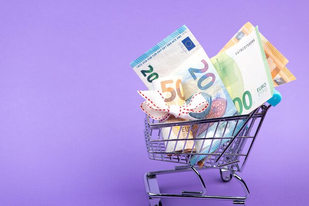 Rouleau de billets en euros avec ruban blanc dans un chariot de supermarché isolé sur fond violet