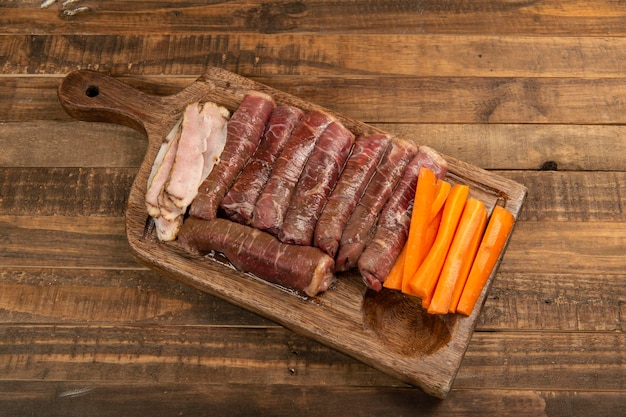 Rouleau de boeuf cru à préparer avec du bacon et des carottes sur une planche de cuisine