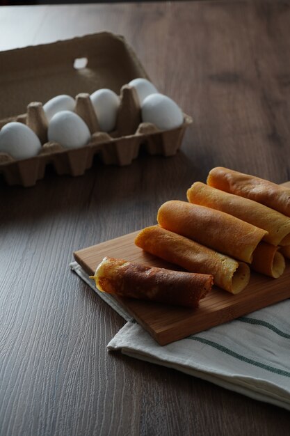 Rouleau de crêpes rempli de saveurs assorties sur une planche à découper en bambou placé sur un tissu rayé blanc et vert sur une table en bois et un paquet d'oeufs