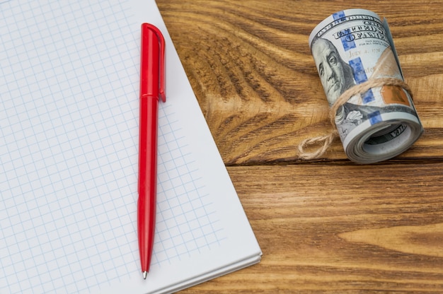 Un rouleau de dollars, un cahier et un stylo se trouvent sur une table texturée en bois.